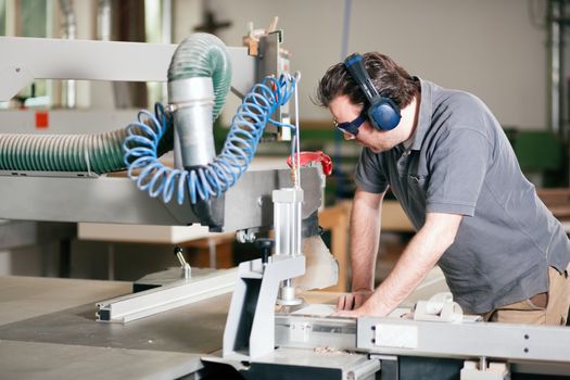 Carpenter working on an electric buzz saw cutting some boards, he is wearing safety glasses and hearing protection to make things safe