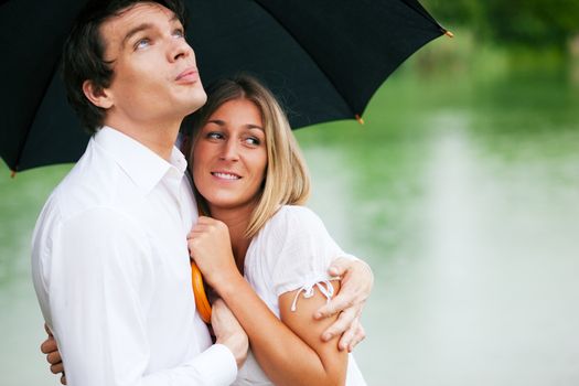 Couple (man and woman) at a lake in summer rain with an umbrella, he is sheltering her from the drops, holding his girl in his arms