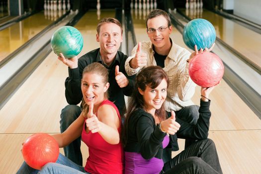 Group of four friends in a bowling alley having fun, holding their bowling balls and showing thumbs up (focus on guys in second row)