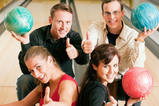 Group of four friends in a bowling alley having fun, holding their bowling balls and showing thumbs up (focus on girls in front row)