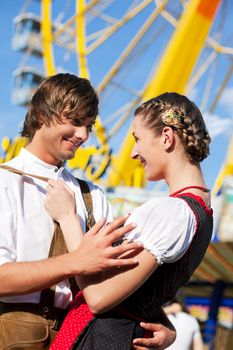 Young and beautiful couple in traditional Bavarian Tracht - Dirndl and Lederhosen - embracing each other on a fair like a Dult or the Oktoberfest; both are standing in front of a big wheel