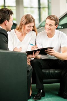 Sales situation in a car dealership, the young couple is signing the sales contract to get the new car in the background