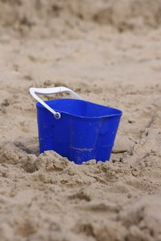 A blue plastic bucket with a white handle set in the sand on a beach.