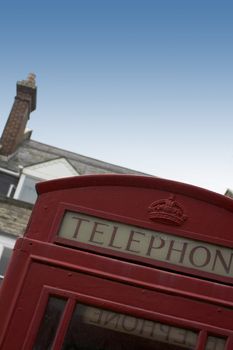 An old English red telephone box. Image set at an angle with roof tops and a chimney visible to the syline background.