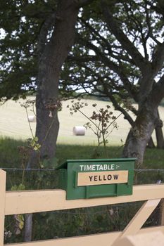 A timetable sign constructed of wood and painted green, set on top of a cream painted fence. Rolling fields with straw bails set to the background. This forms part of the notification system for which timetable is used on the Swanage Steam Railway in Dorset.