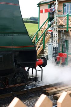 The front end of a period steam engine, letting off steam at the end of the platform at Harmans Cross station, part of the Swanage Steam Railway network in Dorset. The signal box is also visible at the end of the platform.