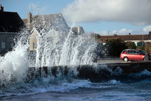 waves splashing against a coastal wall during a storm with cars and homes in background
