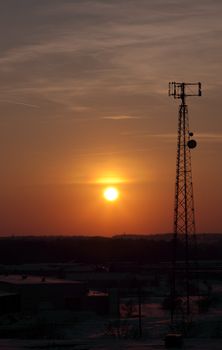 The silhouette of a cell phone tower shot against the orange cast of the setting sun.
