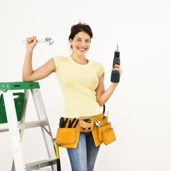 Woman standing in home with ladder and holding tools smiling.