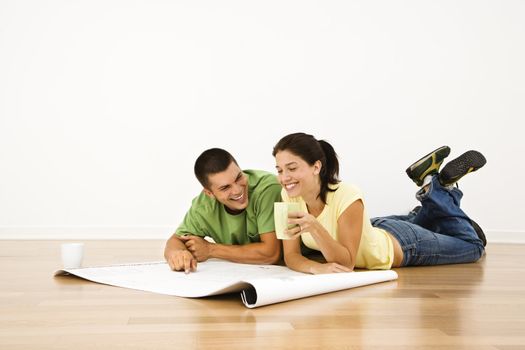 Attractive young adult couple lying on home floor with coffee cups smiling and looking at blueprints.