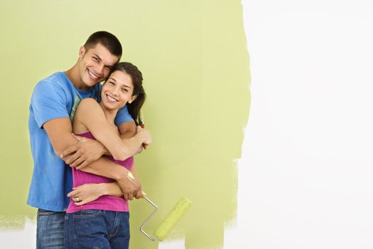 Attractive smiling couple posing in front of partially painted wall holding paint roller.