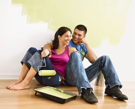 Couple sitting on floor smiling in front of partially painted wall in home.