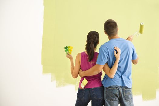 Attractive couple standing in front of partially painted wall with arms around eachother.