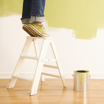Legs of woman standing on tiptoe on stepladder with paint can and painted wall.