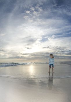 A child watches the surf flow in while the setting sun reflects off the water and the clouds glow on the horizon