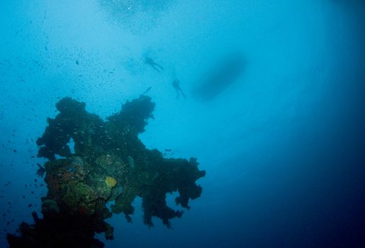 Two divers and a boat silhouetted on the surface above a coral encrusted mast from a World War II shipwreck in Truk Lagoon, Micronesia