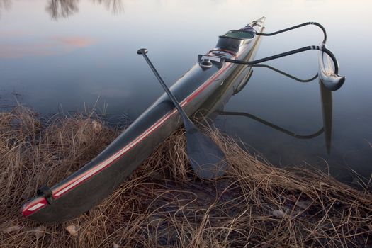 long and slim racing outrigger canoe (black carbon fiber design) with a paddle on shore of calm lake