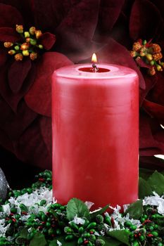 Burning Christmas candle surrounded by rich burgundy colored Poinsettias, holly and snow.
