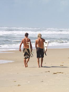 Two young male surfer dudes walking along the beach away from the camera carrying their surfboards with the waves crashing in the background