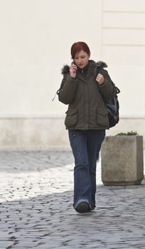 Redheaded girl student at the phone while is walking on a paved street.