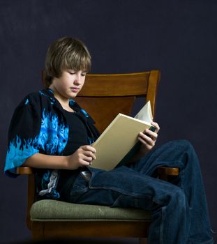 A young teenage boy reading a book seated in an armchair.