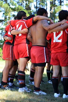 Rugby team with players in red jerseys standing together by holding their arms around each other before a training session for an important game on the field outdoors