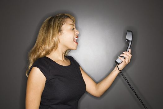 Attractive woman yelling into phone receiver standing against gray background.