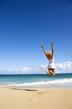 Pretty young woman jumping on Maui, Hawaii beach with arms raised into air.