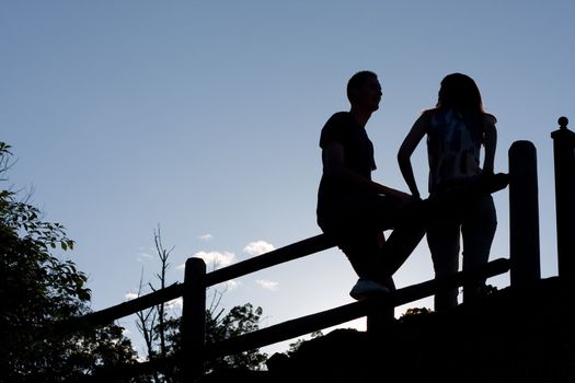 Silhouette of a young couple hanging out together outdoors by an old country fence during the early evening hours.