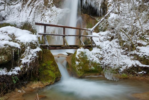 Waterfall with icicles and snow around with a small wooden bridge on a winter day.