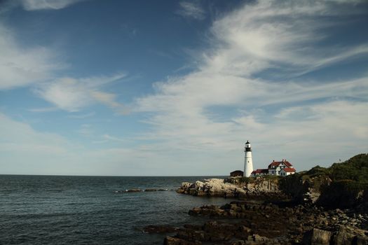 Portland Haed Lightouse in the distance on a rocky clif with partly cloudy skies.