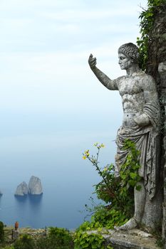 A Roman statue with its arms raised and rocks rising from the sea in the background.
