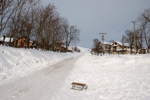 Hill road covered with snow going between two rows of houses with old sled left in foreground.