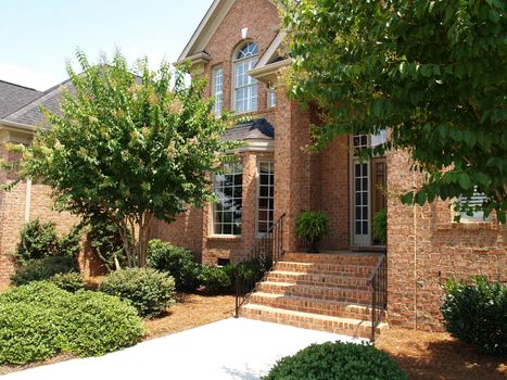 Front of a large brick home with arched windows, beautiful landscaping and wrought iron railings on the steps. 