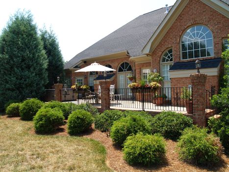 Large brick patio with an iron railing on the back of an expensive home with arched windows. Well-kept planters and a covered table decorate the exterior room.