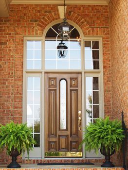 Front door of a brick luxury home with a wooden door, hanging light and planters with ferns.