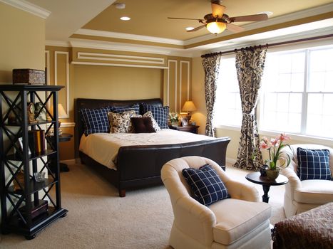 Nicely decorated master bedroom in a newly built luxury home. There is a black leather sleigh bed, dark furniture, light streaming through the windows, and a light sitting area.