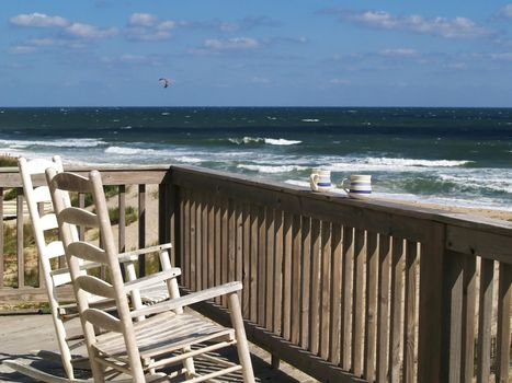 Two rocking chairs and a couple of coffee mugs on a deck overlooking a beach scene. 