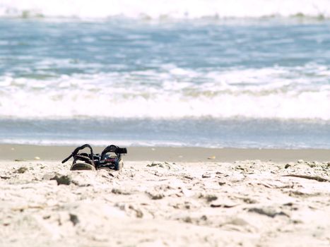 A pair of dark sandals sitting alone on a sandy beach with the surf in the background and a narrow depth of field