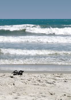 A pair of dark sandals sitting alone on a sandy beach with the surf in the background and a narrow depth of field