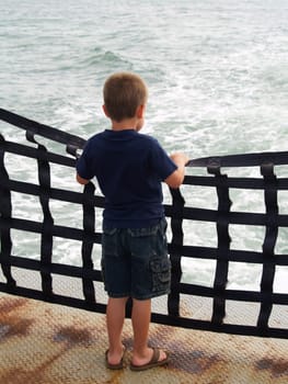 Young boy in jeans shorts and a blue shirt and sandals on the back of a passenger ferry watching the churning water while holding onto the webbed railing.