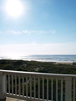 View of a bright pristine sunlit summertime beach from an upper deck with the white painted railing and a bit of the deck visible.