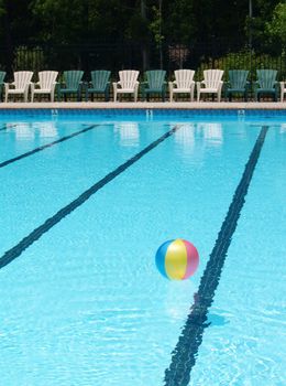 A beach ball floating on the surface of a refeshing swimming pool in the summertime with a row of chairs on the deck. Let's go to the pool!