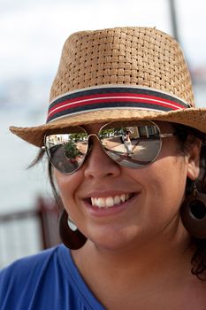An attractive young Hispanic woman smiling with her hat and sunglasses on outdoors.