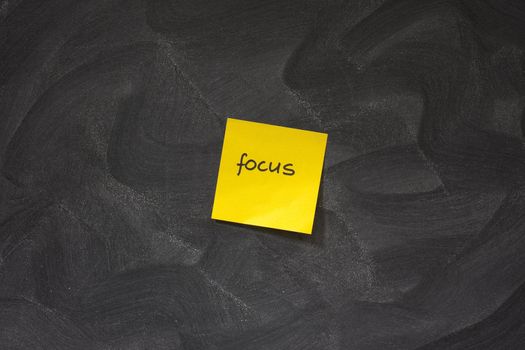 yellow sticky note with word focus posted on blackboard with chaotic white chalk eraser patterns