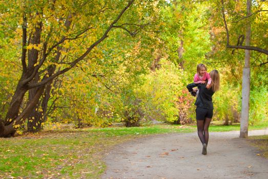 Mom is carrying away her daughter (3 years old). They are walking along the road in autumn park