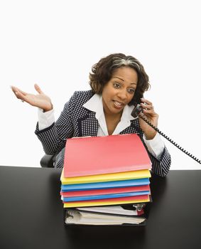 Businesswoman sitting at office desk with large stack of work talking on telephone and gesturing.