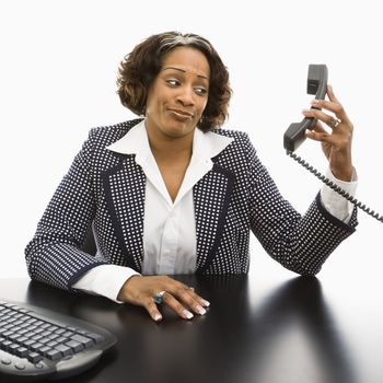 Businesswoman sitting at desk holding telephone receiver out away from head looking with strange expression.
