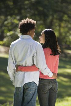 Rear view of couple with arms around eachother looking into eachother's eyes in park.