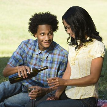 Man pouring woman glass of wine sitting on picnic blanket in park.